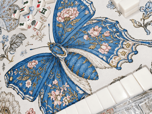 Close-up of the Blue Butterfly Garden Mahjong Mat showing a blue butterfly with pink floral details, textured ivory fabric, and mahjong tiles along the edges.