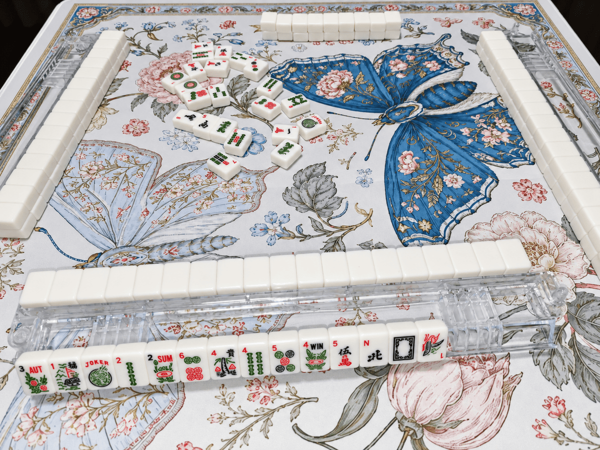 Angled view of the Blue Butterfly Garden Mahjong Mat with blue butterflies, pink flowers, and an ornate border, arranged with ivory mahjong tiles and clear racks.