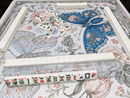 Angled view of the Blue Butterfly Garden Mahjong Mat with blue butterflies, pink flowers, and an ornate border, arranged with ivory mahjong tiles and clear racks.