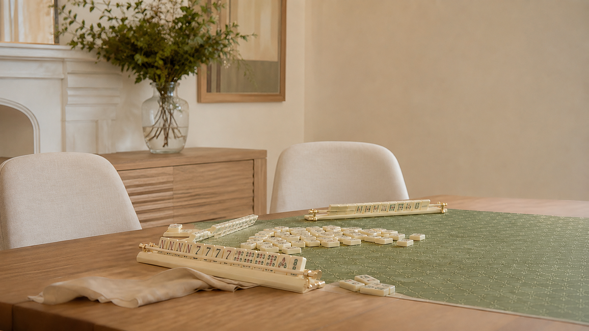 Ivory mahjong tiles and gold-accented racks on a patterned sage green mat atop a wooden table.