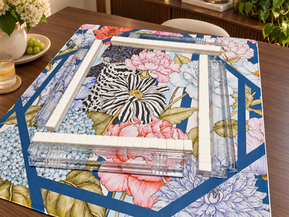 Black leopard floral mahjong mat with ivory tiles and clear racks on a walnut table in a sunlit room with flowers and greenery.