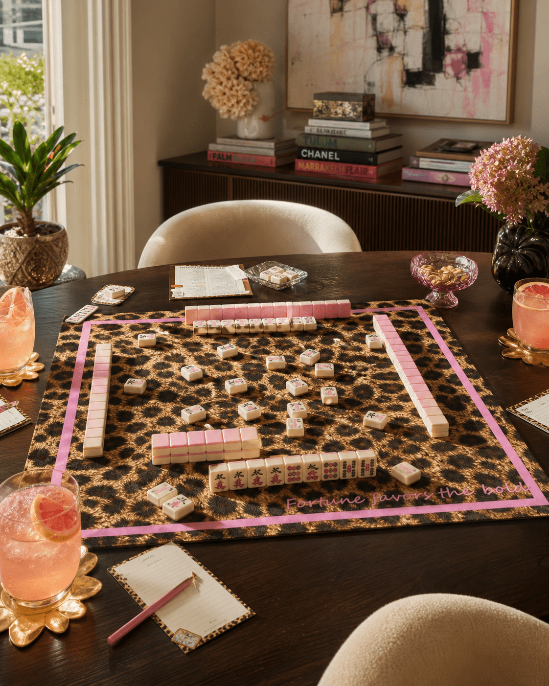 Leopard-print mahjong mat with a pink border arranged with tiles, racks, dice, and drinks on a dark table.