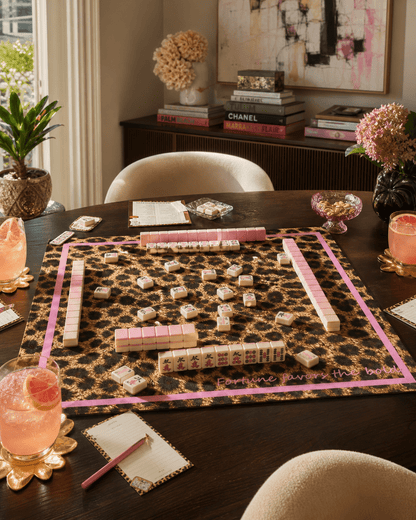 Leopard-print mahjong mat with a pink border arranged with tiles, racks, dice, and drinks on a dark table.