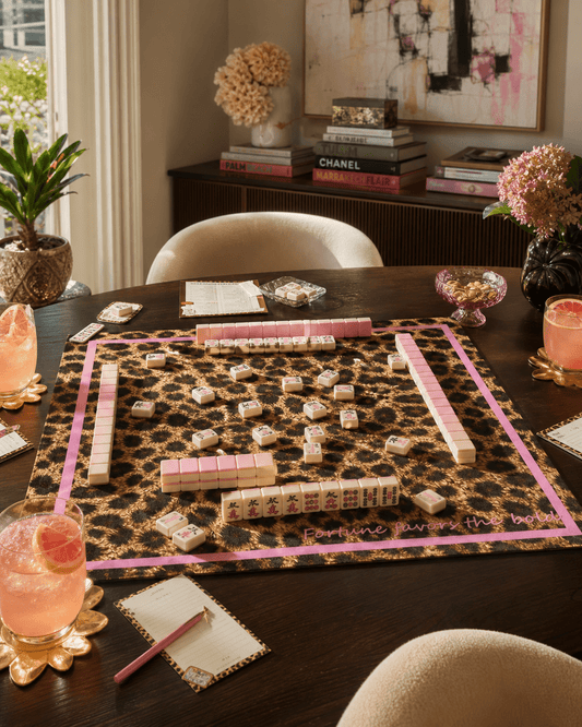 Leopard-print mahjong mat with a pink border arranged with tiles, racks, dice, and drinks on a dark table.