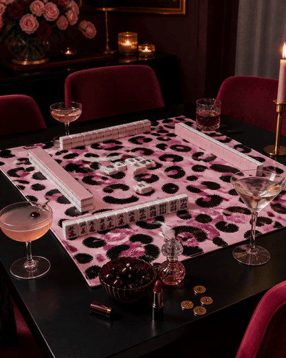 Pink leopard-print mahjong mat set for play on a dark table in a glamorous candlelit room.